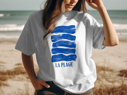 White t-shirt with blue brushstroke design and "LA PLAGE" text on a beach.