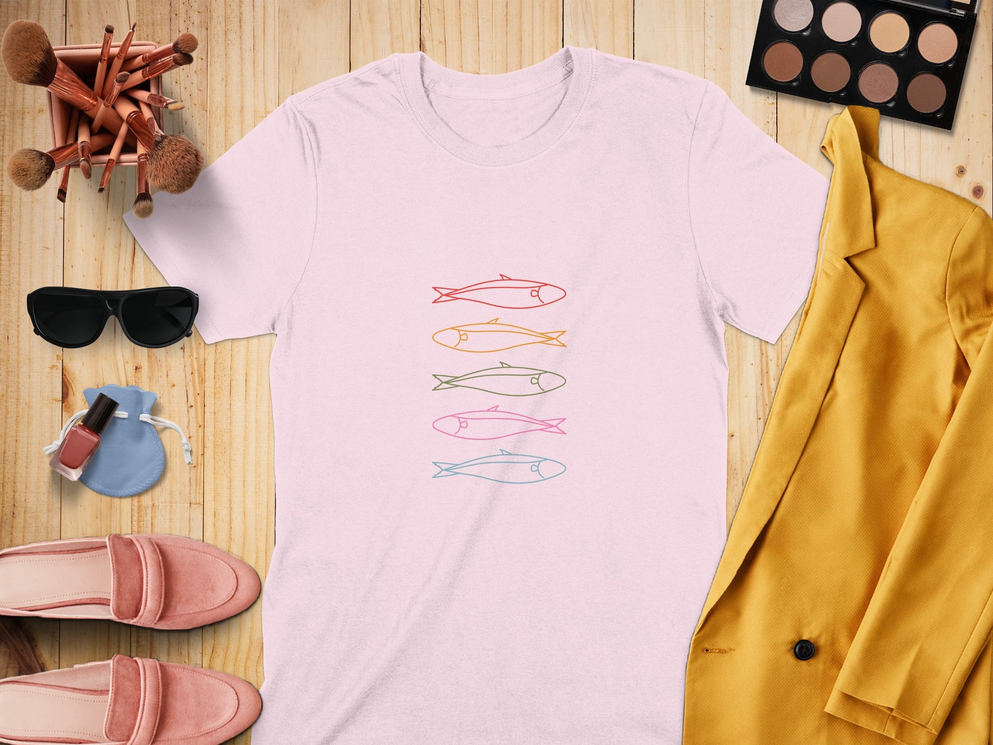 Pink t-shirt with five colorful cartoon fish illustration, flat lay with makeup and fashion items.