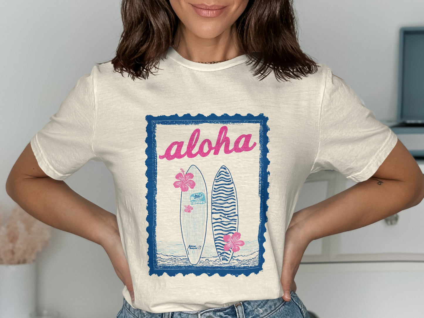 Cream t-shirt with blue stamp graphic featuring surfboards, hibiscus flowers, and "Aloha" text.