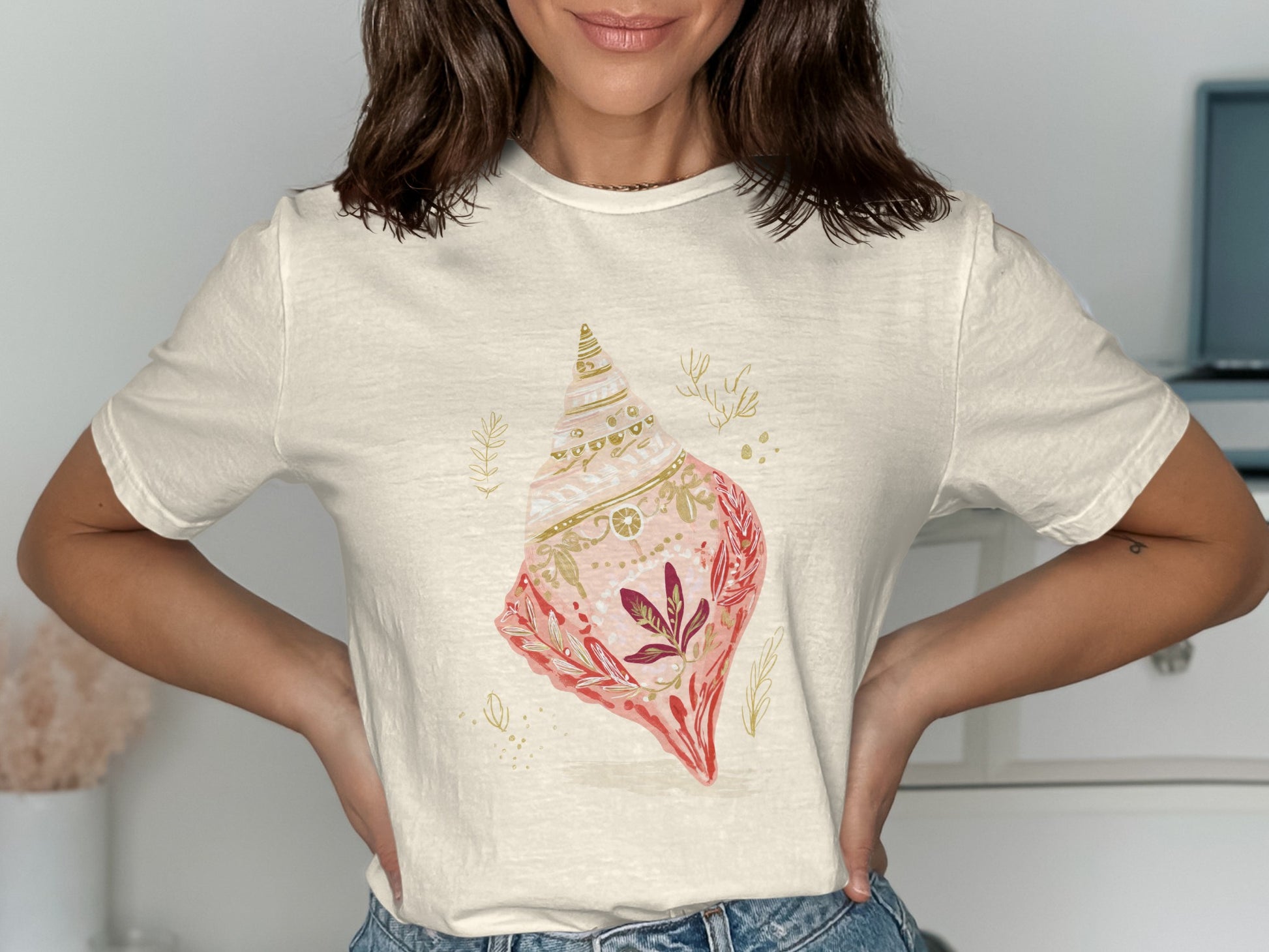 Cream t-shirt with a watercolor seashell graphic featuring pink and gold accents.