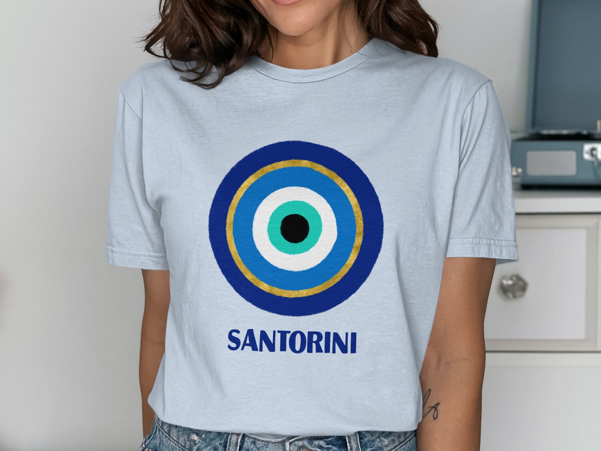 Light blue t-shirt with a blue and gold evil eye design and "SANTORINI" text.