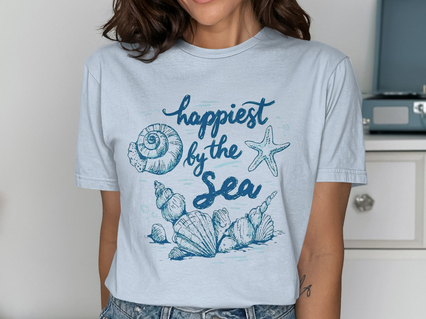 Light blue t-shirt with seashells and "happiest by the sea" text.