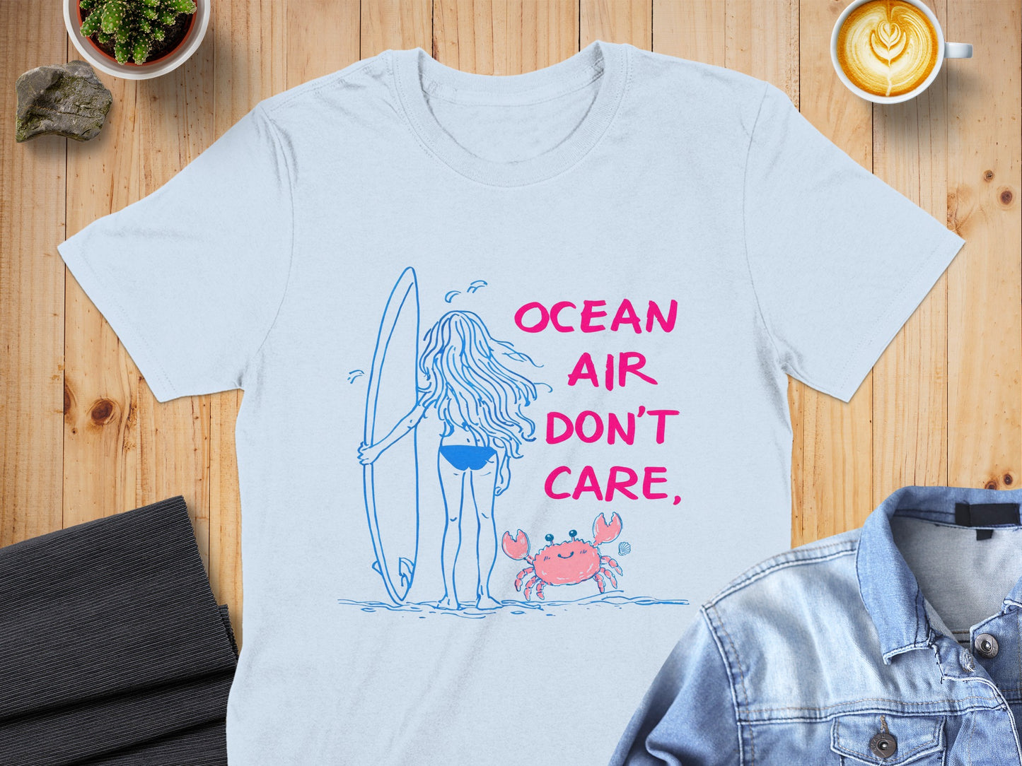Blue t-shirt with a graphic of a person with a surfboard and a crab, reading "Ocean Air Don't Care".