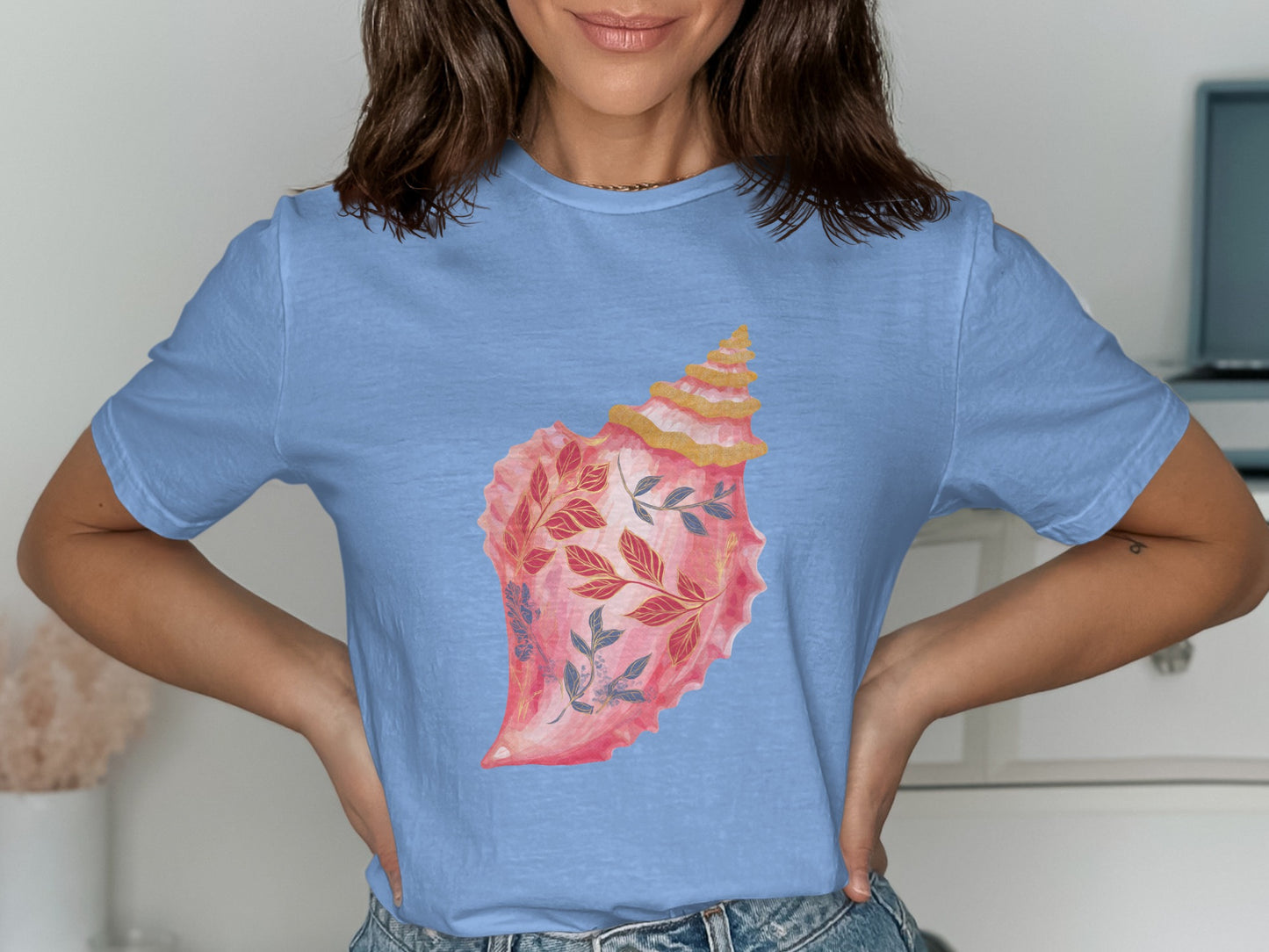 Light blue t-shirt with a watercolor illustration of a pink seashell adorned with floral branches.