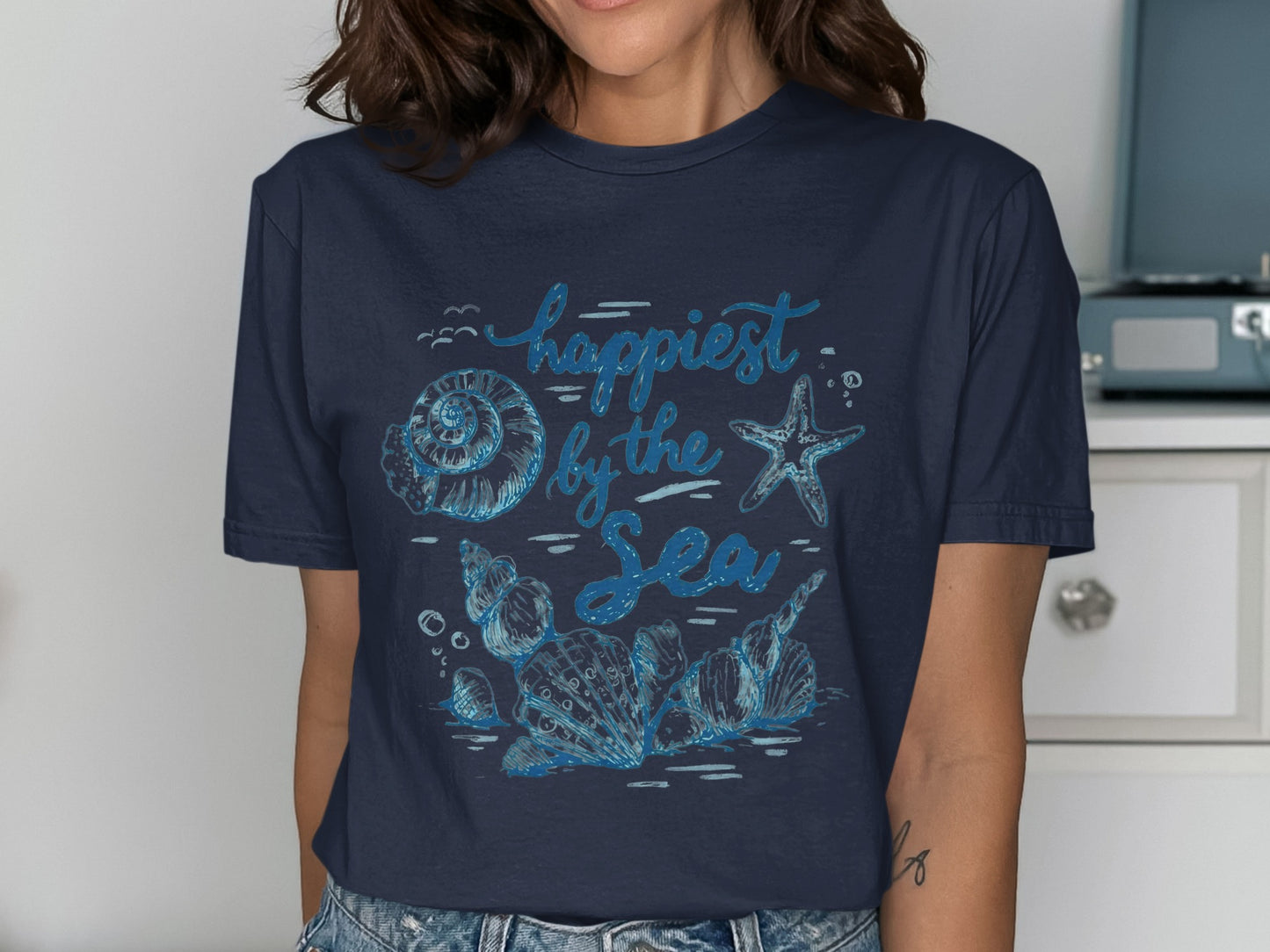 Navy t-shirt with blue "Happiest by the Sea" text and seashell illustrations.