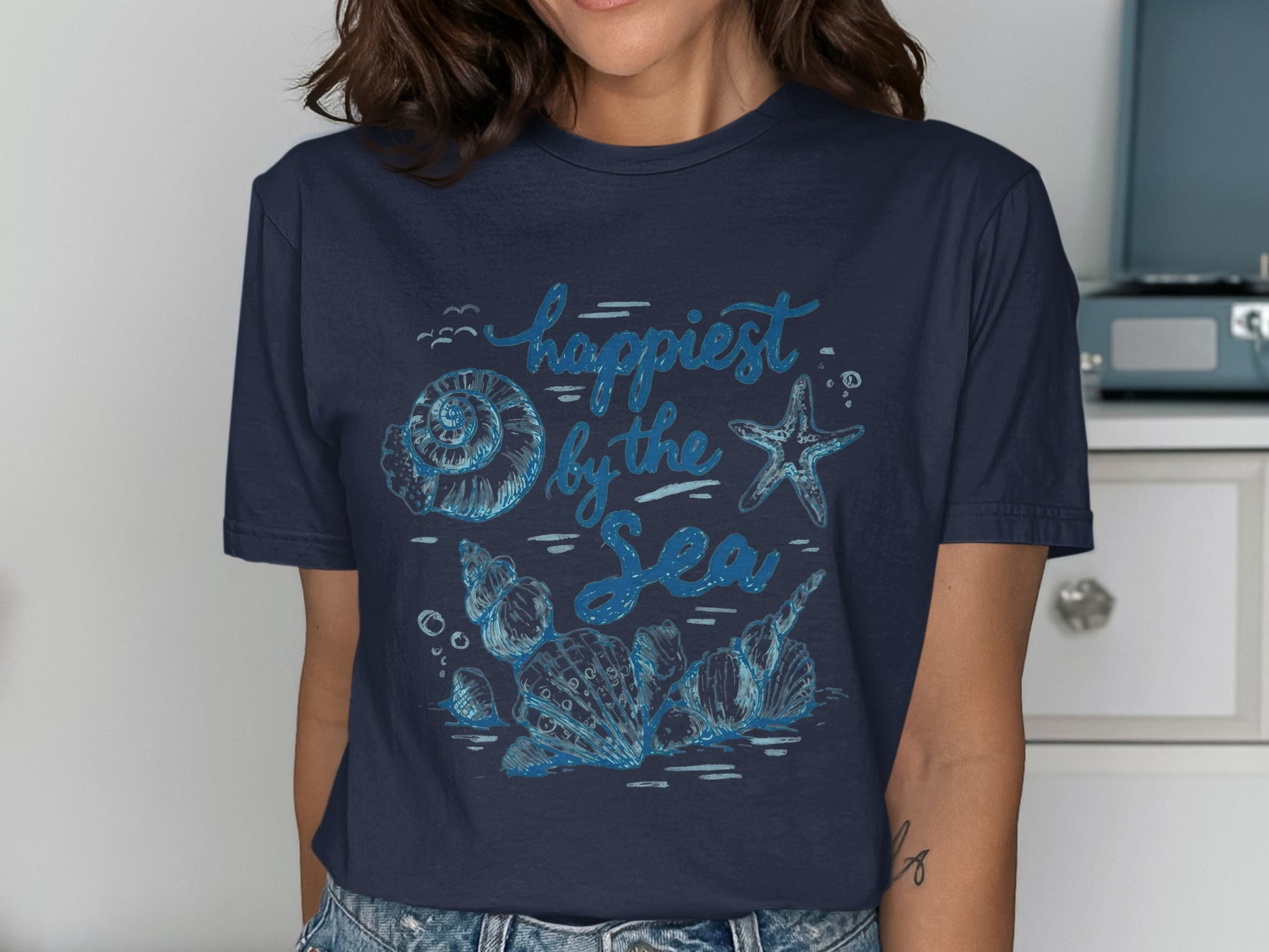 Navy t-shirt with blue "Happiest by the Sea" text and seashell illustrations.
