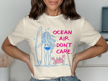 Cream t-shirt with a woman holding a surfboard and a crab, reads "OCEAN AIR DON'T CARE".