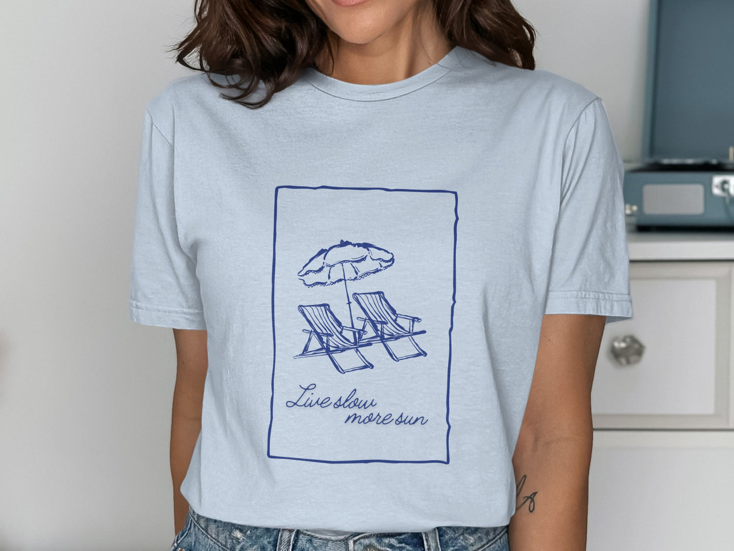 Light blue t-shirt with drawing of two lounge chairs under an umbrella and text "Live slow more sun".