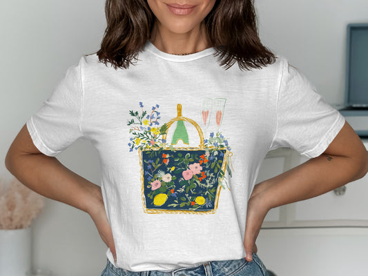 White t-shirt with a graphic of a picnic basket with flowers, lemons, and champagne.
