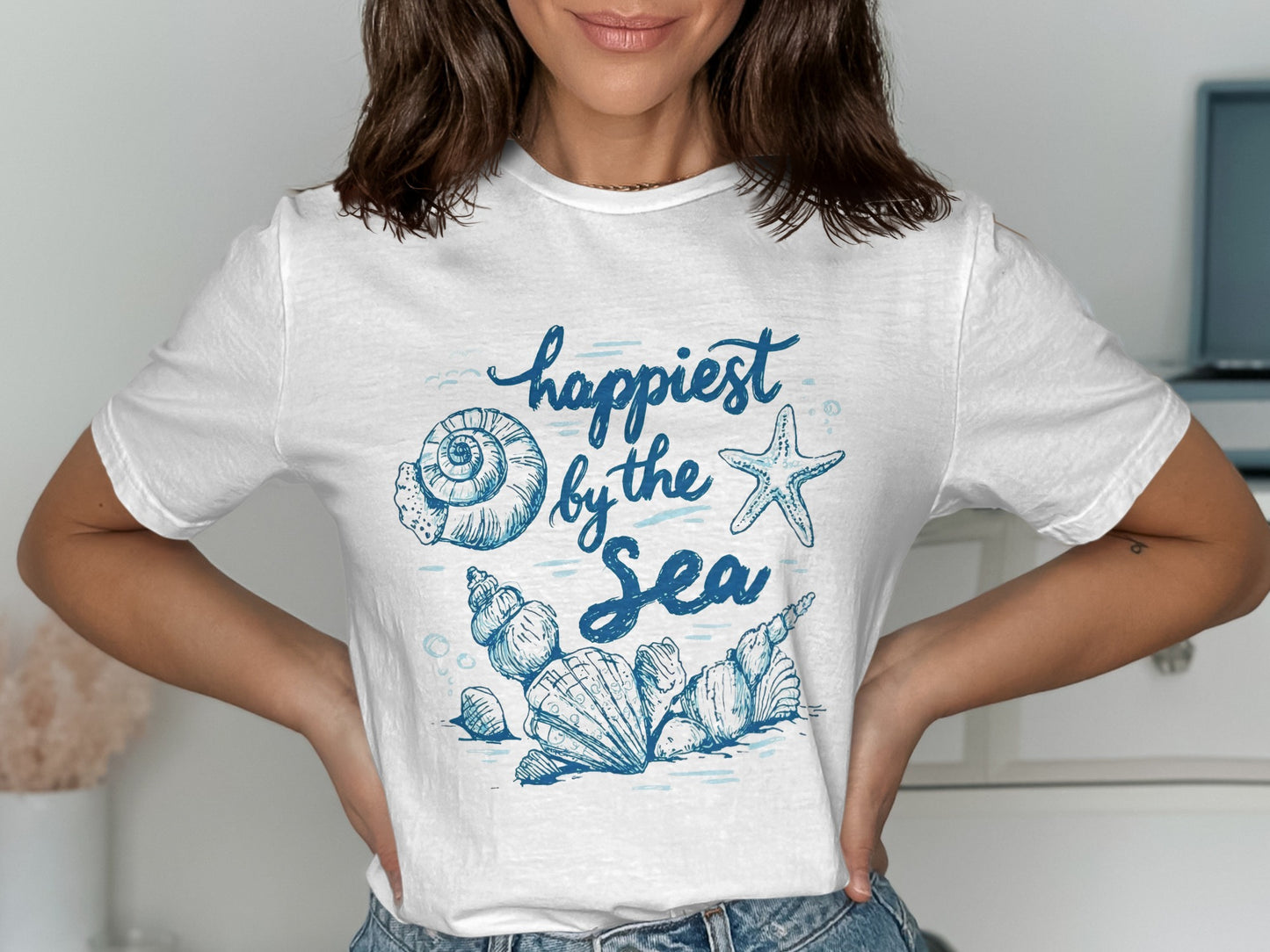 White t-shirt with blue "Happiest by the Sea" text and ocean shell graphics.