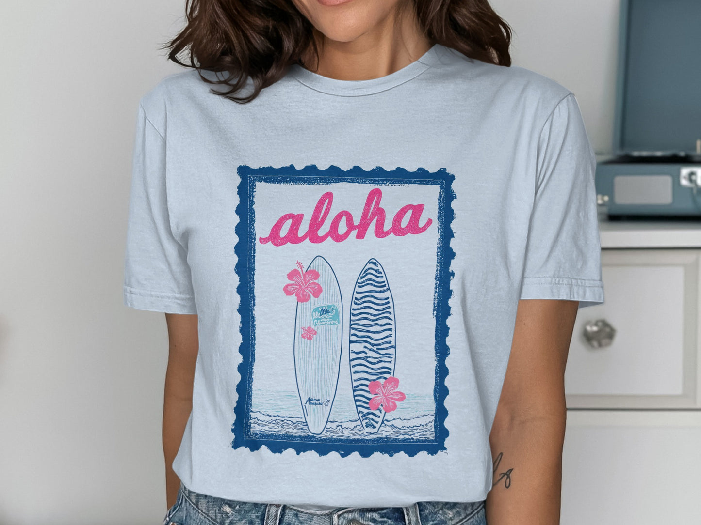 Light blue t-shirt with graphic of two surfboards, flowers, and "aloha" text.