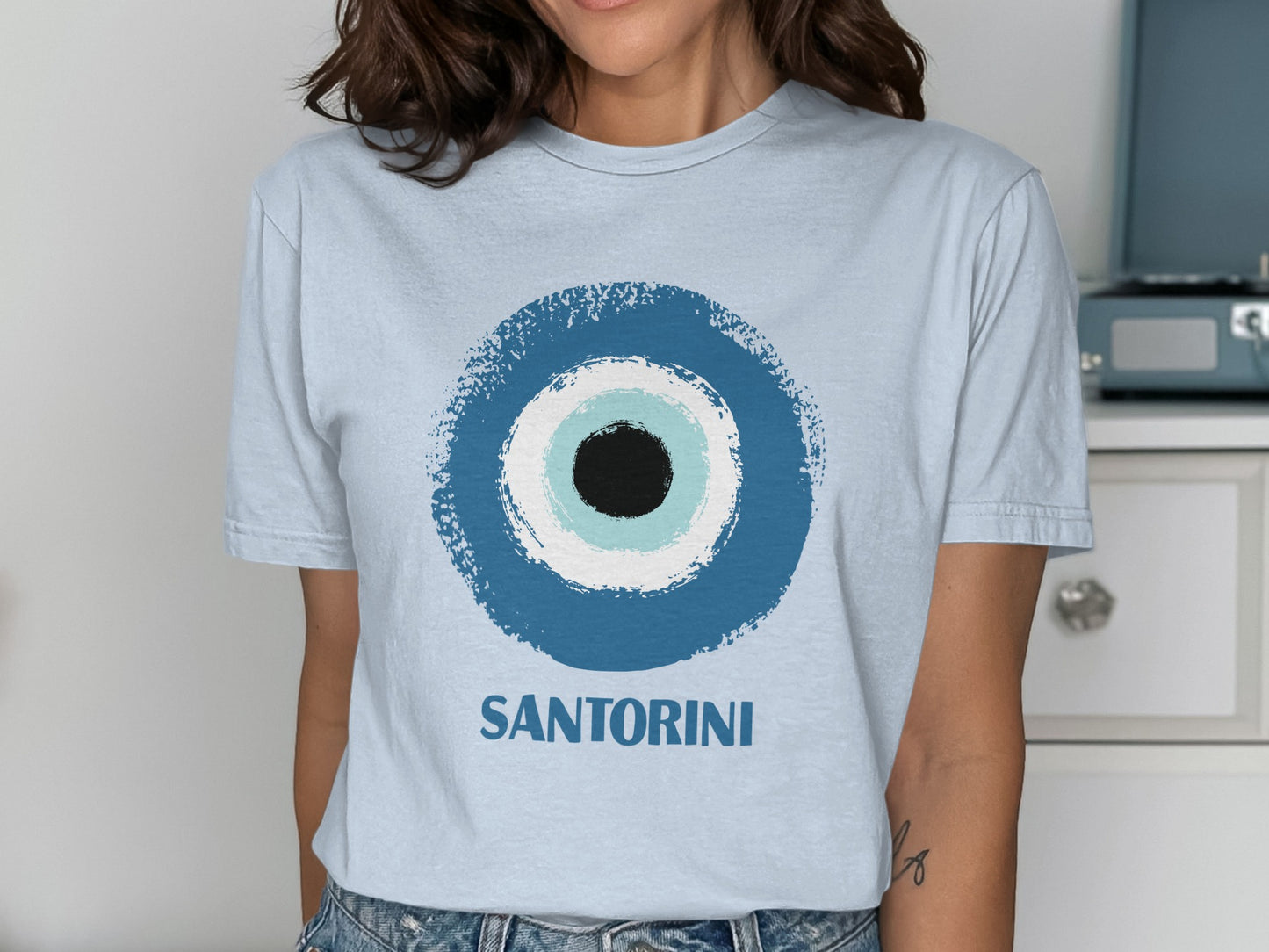 Light blue t-shirt with a blue, white, and black evil eye design and "SANTORINI" text.
