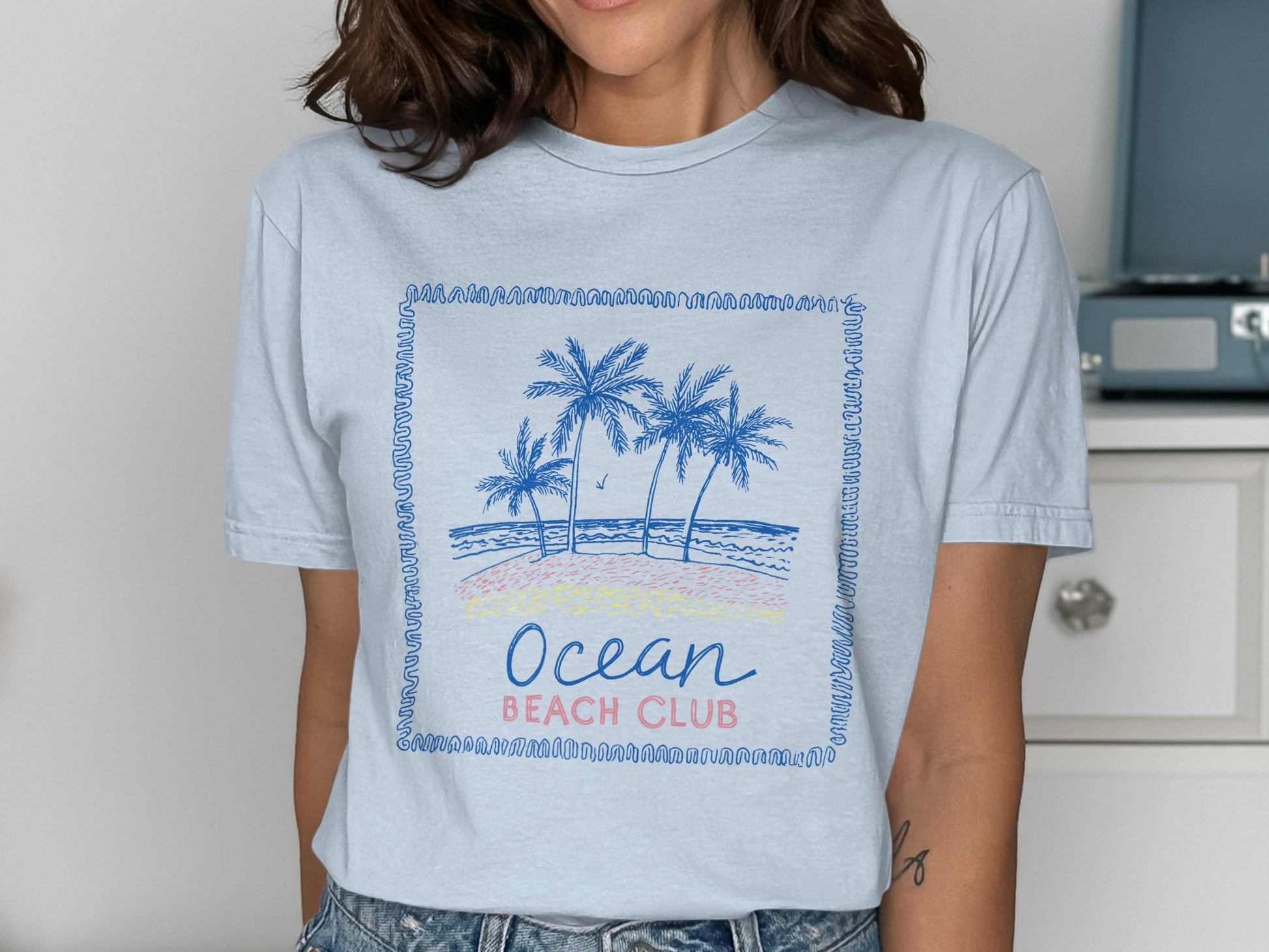 Light blue t-shirt with a framed beach scene and "Ocean Beach Club" text.