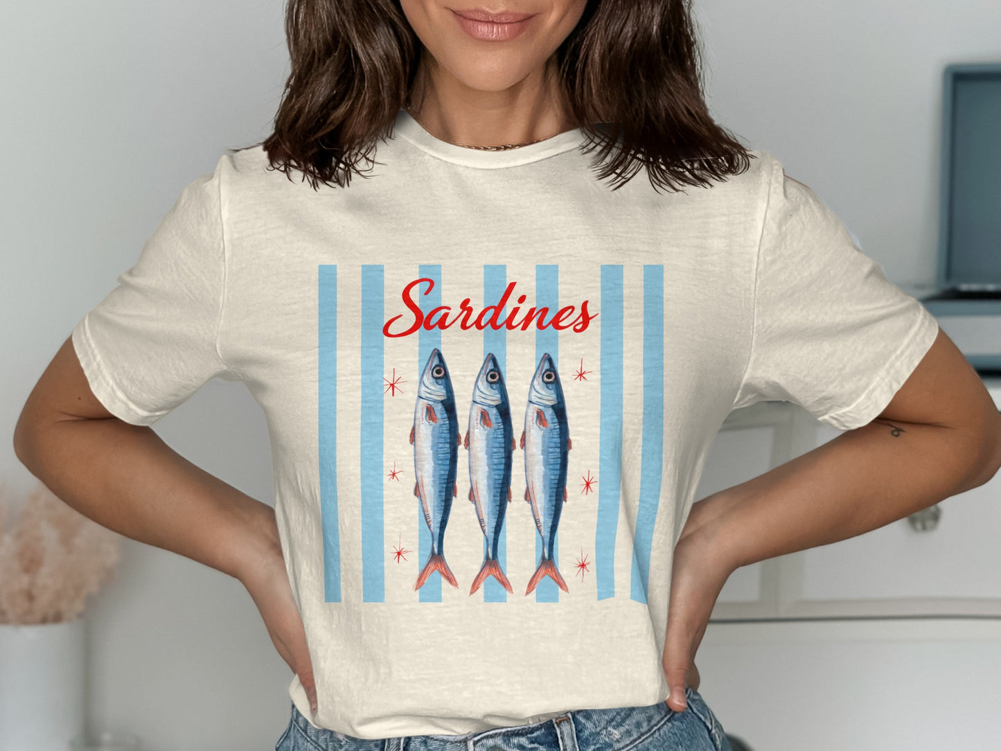 Cream t-shirt with three stylized sardines and red text "Sardines" on a blue striped background.
