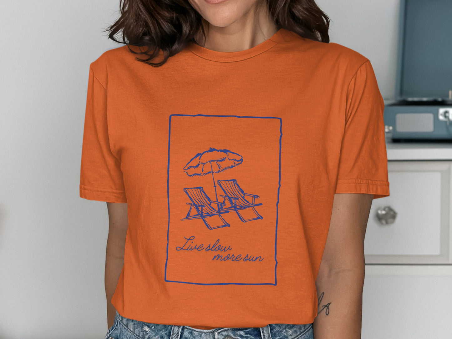 Orange t-shirt with a blue line drawing of beach chairs and umbrella, with text "Live slow, more sun."