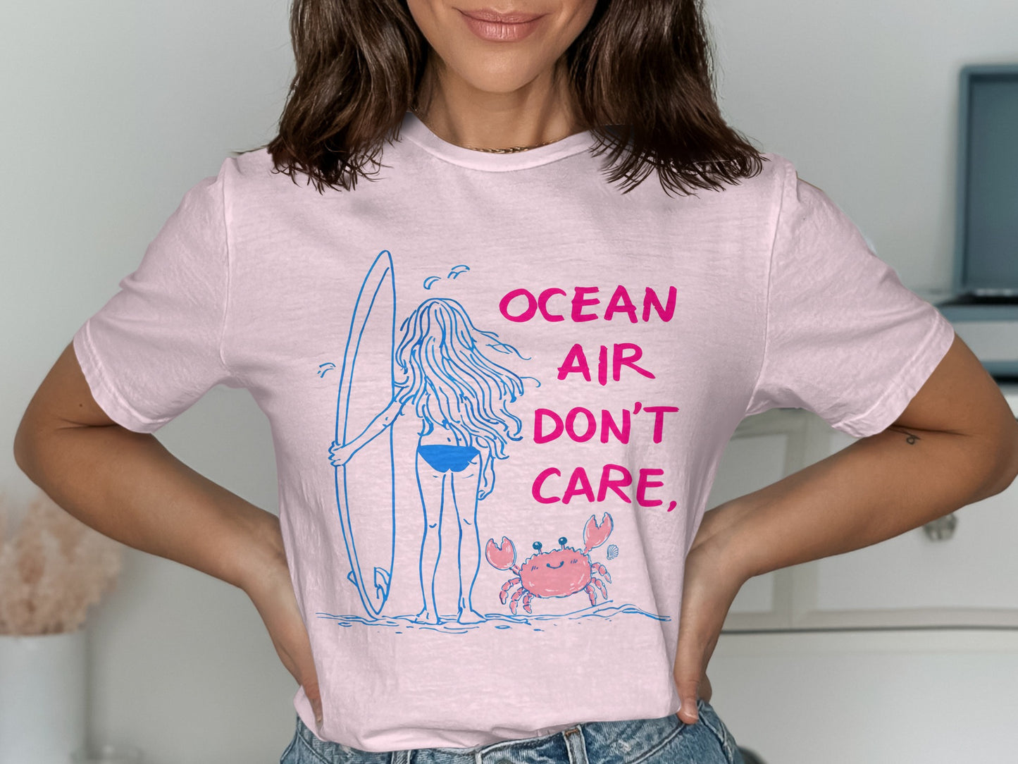 Pink t-shirt with a drawing of a girl holding a surfboard and a crab, text reads "OCEAN AIR DON'T CARE."