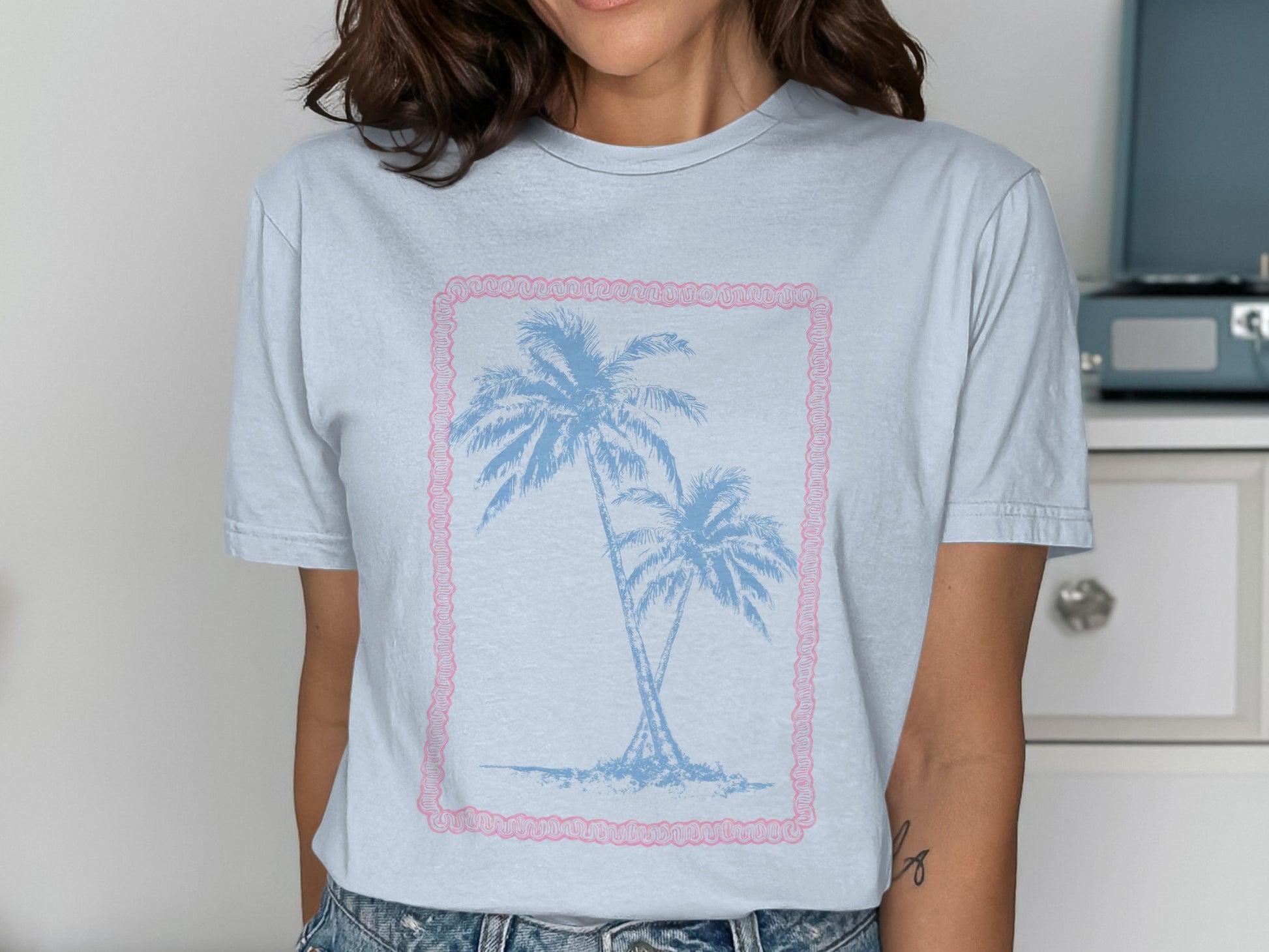 Light blue t-shirt with two palm trees inside a pink frame.