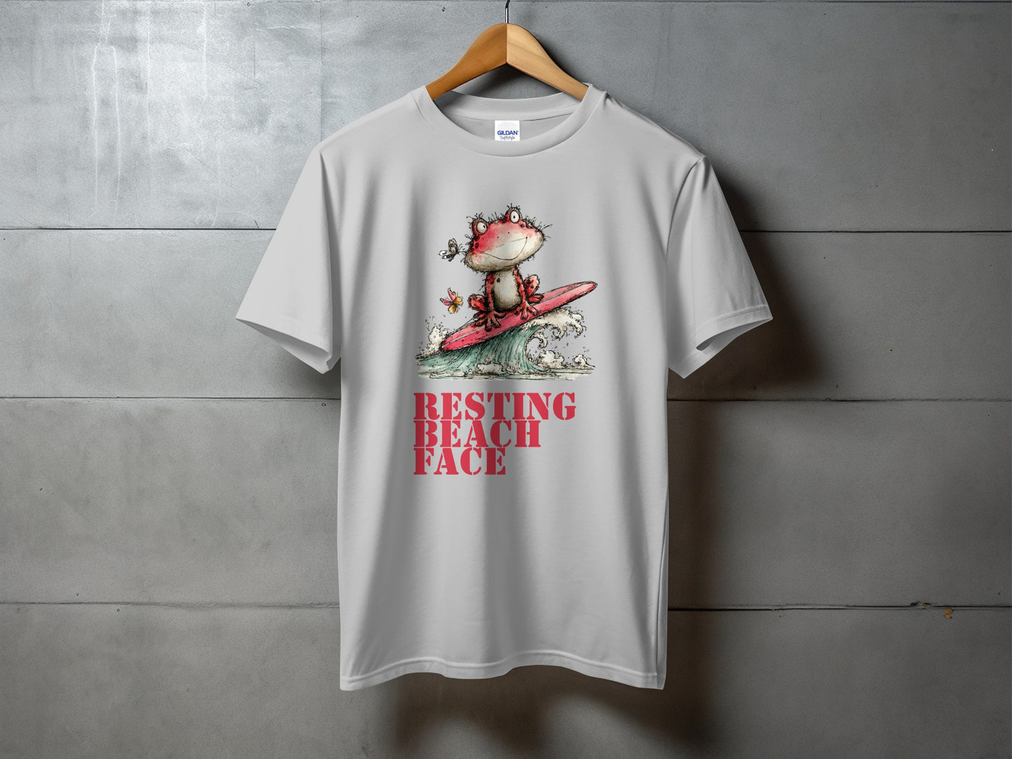 White t-shirt with graphic of frog surfing and text "RESTING BEACH FACE."