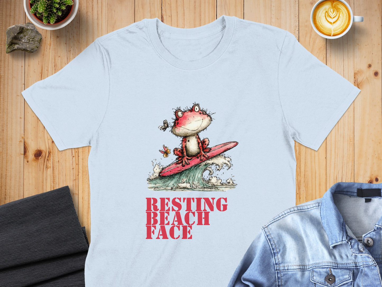 Light blue t-shirt with a cartoon frog surfing a wave, text reads "RESTING BEACH FACE".