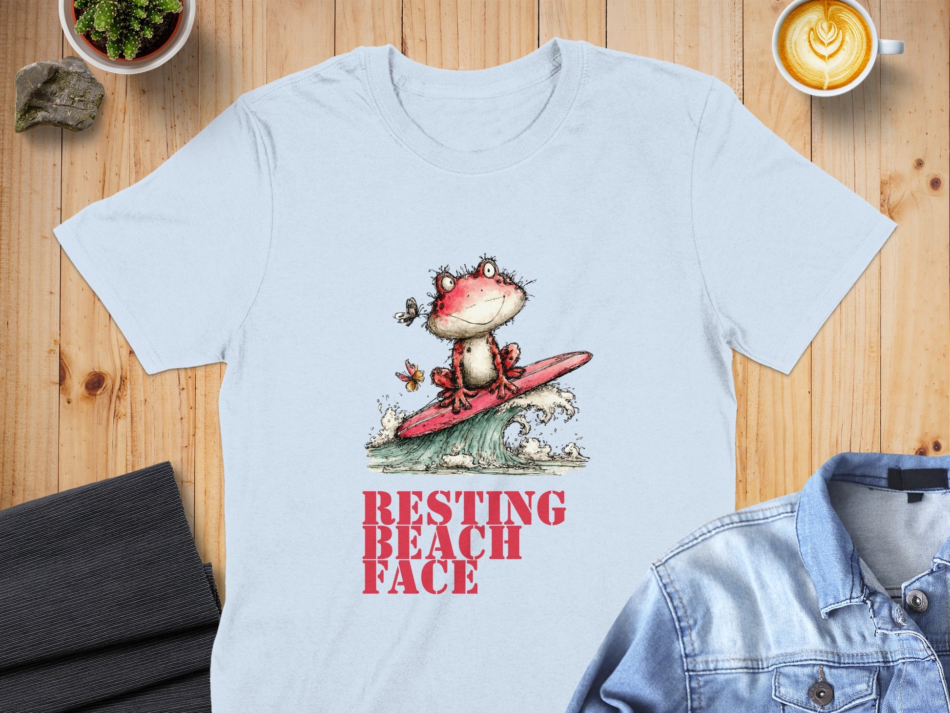 Light blue t-shirt with a cartoon frog surfing a wave, text reads "RESTING BEACH FACE".