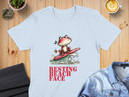 Light blue t-shirt with a cartoon frog surfing a wave, text reads "RESTING BEACH FACE".