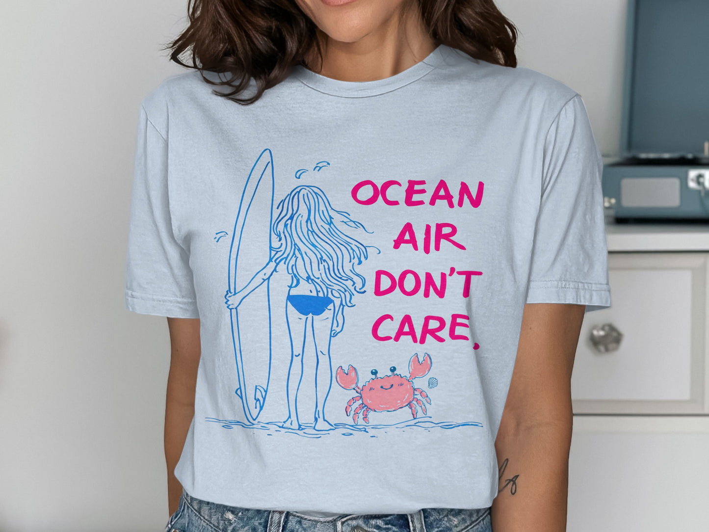 T-shirt with a drawing of a surfer girl and a crab, text reads "OCEAN AIR DON'T CARE".