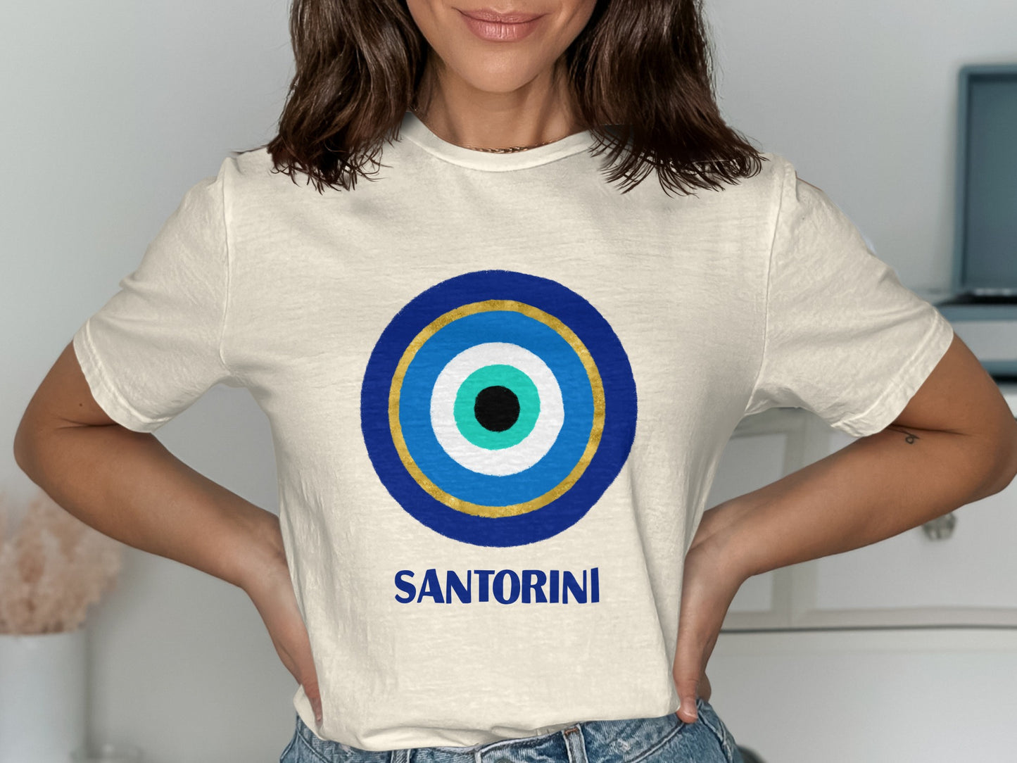 Cream t-shirt with a blue, gold, and white evil eye design and "SANTORINI" text.