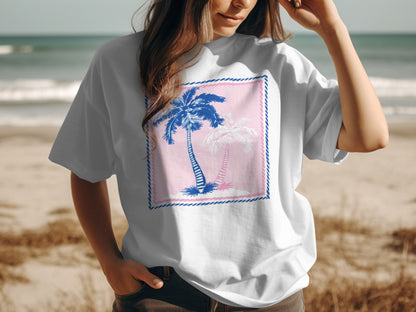White t-shirt with pink and blue palm tree graphic on a beach.