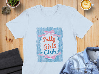 Light blue t-shirt with "Salty Girls Club" text and pink bows on a wooden surface.