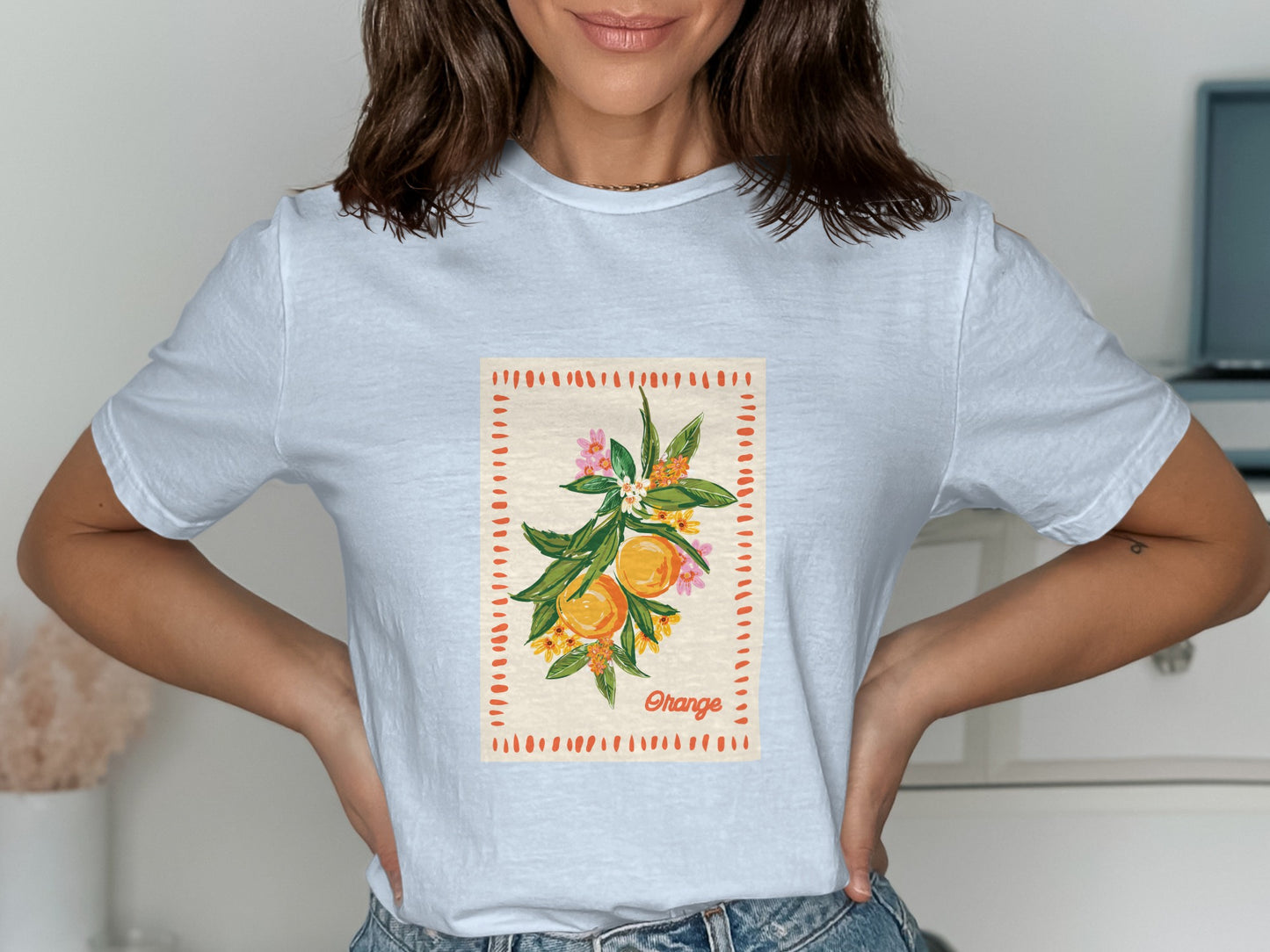 White t-shirt with orange branch and fruit design, framed by dashed lines and small flowers.