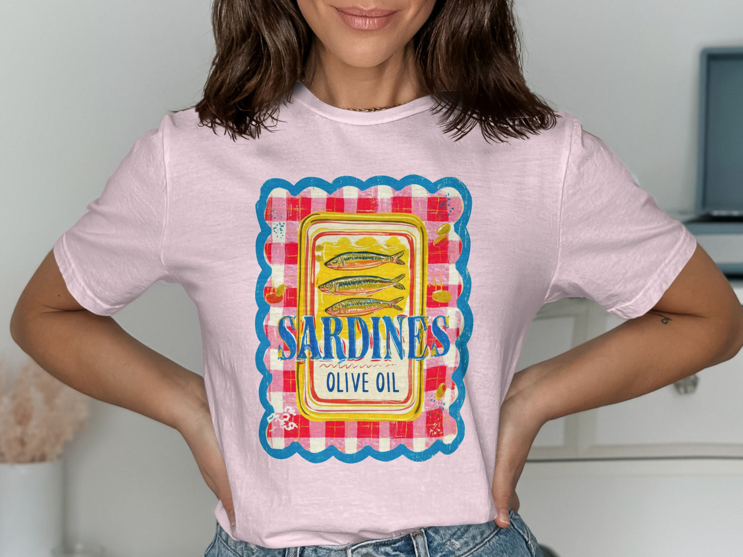 Pink t-shirt featuring a graphic of sardines in olive oil within a gingham border.